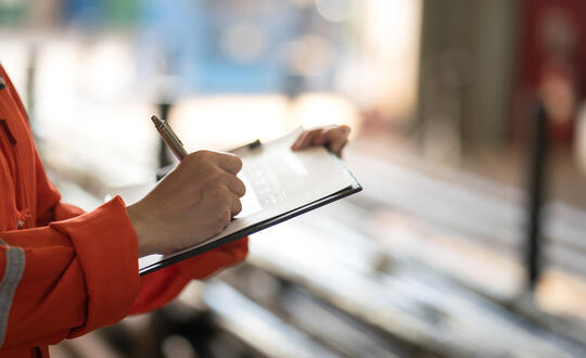 A construction worker in high-visibility clothing conducting a safety inspection and taking notes on a clipboard at a worksite.
