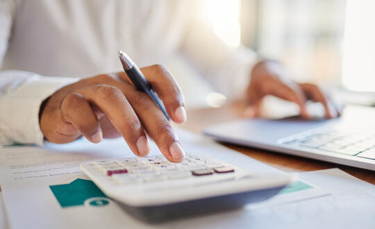 Close-up of a professional working with financial documents, using a calculator and laptop on a wooden desk in a sunny office environment.