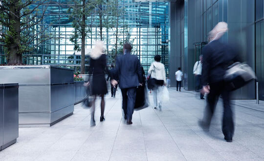 Commuters walking briskly in front of a modern glass facade office building, capturing the hustle and bustle of city life during rush hour.