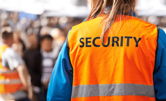 A security personnel wearing a high-visibility orange jacket with SECURITY emblazoned on the back, overseeing a crowded event.