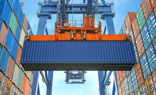 Gantry crane lifting a blue shipping container amidst stacked colourful containers at a commercial cargo port under a clear sky.