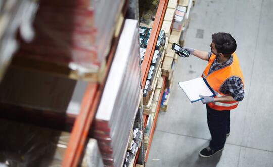 Warehouse worker in high visibility vest conducting inventory check in industrial storage area.