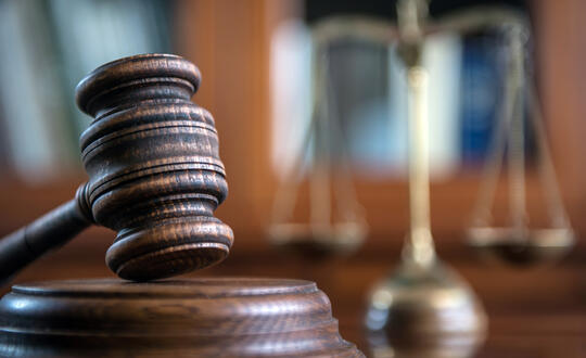 Close-up of a wooden judge's gavel on a desk with a blurred background featuring the scales of justice, symbolising legal authority, courtroom proceedings, and the judicial system in the UK.