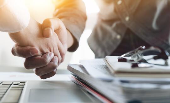 Two professionals engaging in a handshake over a business desk, symbolising partnership, collaboration, and agreement with laptops and paperwork in the background, in an office environment.