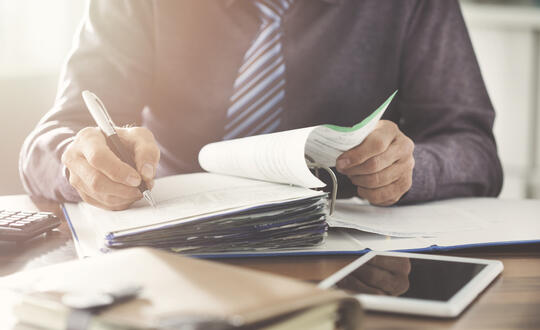 Business professionals reviewing financial documents and making notes, with focus on hands writing in a ledger on a cluttered office desk, including a tablet and calculator.