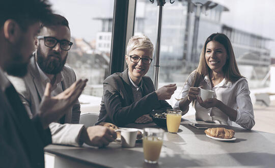 Group of cheerful colleagues enjoying a casual business breakfast at a sunlit café, engaging in conversation over fresh orange juice and croissants.