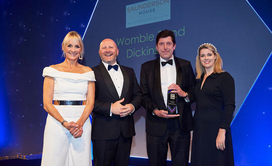 Four professionals posing with an award at a formal evening event, with the Womble Bond Dickinson logo in the background