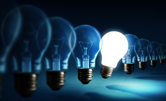 A row of unlit incandescent bulbs with a single glowing bulb standing out, symbolising innovation and creative ideas, set against a dark blue background.