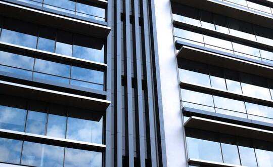 Modern commercial building facade with reflective glass windows and steel structure against a clear blue sky in an urban setting.
