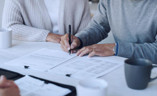 Two individuals collaborating on paperwork, signing documents in a business meeting environment, with coffee mugs on the table.
