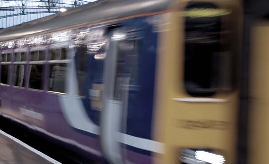 Blurred image of a moving train at a railway station, showcasing motion blur, with a focus on the modern purple and gold livery of a British passenger train under a glass canopy.