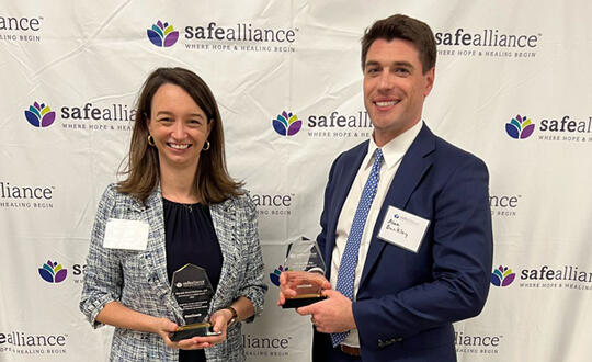 Sarah Motley Stone and Alex Buckley stand in front of a Safe Alliance banner, holding awards in recognition of Buckley's pro bono work and the firm's ongoing commitment to Safe Alliance.