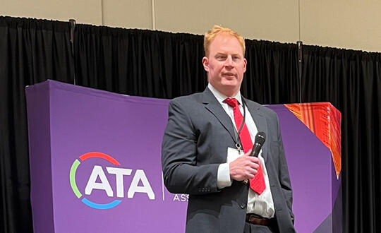 Attorney Cory Schug holds a microphone in front of a purple banner reading "ATA" for the Advanced Textiles Association.