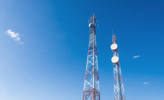 Two towering mobile communication masts against a clear blue sky, equipped with various antennas and satellite dishes for wireless network signal transmission.