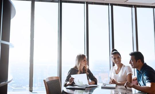Business meeting in a high-rise office with panoramic city views, featuring two women and one man discussing documents