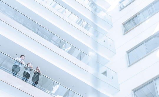 Modern office building interior with bright natural lighting, featuring glass balustrades, clean architectural lines, and a businessperson leaning on the railing of an open-plan walkway.