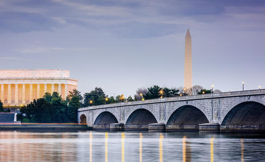 Twilight view over the calm waters of the Potomac River featuring the iconic Lincoln Memorial and Washington Monument in Washington, D.C., with a clear sky and the Arlington Memorial Bridge in the foreground.