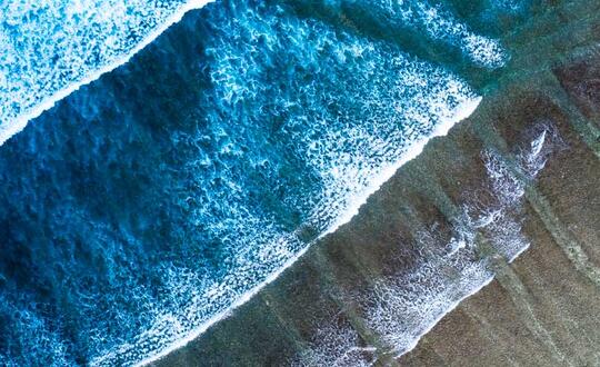 Aerial view of crystal-clear turquoise ocean waves crashing onto a sandy beach with shadows under the water suggesting marine topography.