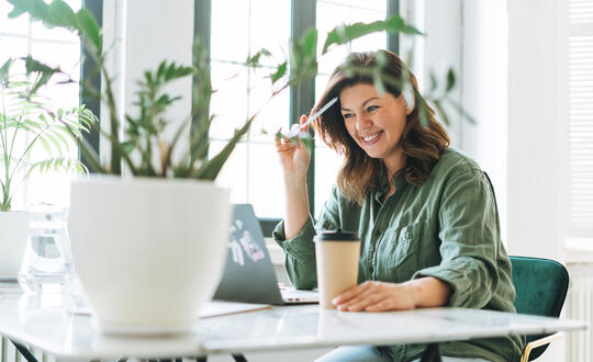 Smiling woman working from home at her minimalistic desk space, with a laptop, coffee cup, and houseplants in a bright, airy room.
