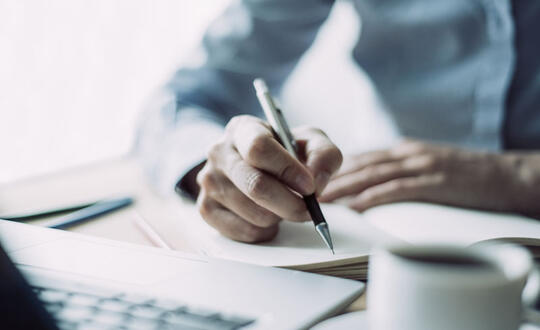 Close-up of a person's hands writing in a notebook with a pen, with a blurred coffee cup in the foreground, suggesting a focus on business planning or creative writing in a relaxed environment.