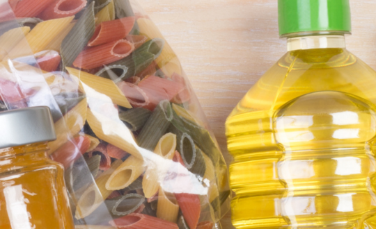 Assortment of staple Italian cooking ingredients including multicoloured pasta, olive oil, and jars of tomato sauce on a wooden worktop.