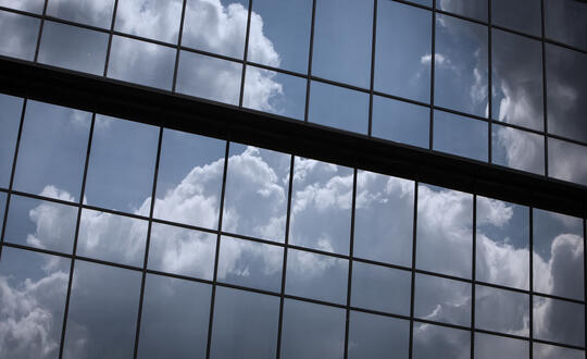 Reflective glass facade of a modern office building with clouds and sky mirrored in the windows, showcasing architectural design and urban landscape.