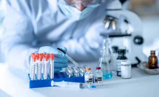 A scientist conducting research in a laboratory, pipetting a sample into test tubes with a rack, surrounded by laboratory equipment and chemical bottles, in a clean and sterile environment.