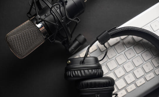 Headphones lay on top of a computer keyboard, while a podcast microphone is positioned beside them.