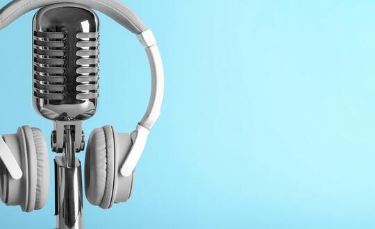 A pair of headphones sit on top of a podcast-style microphone in front of a light blue background.
